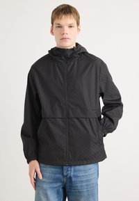 Zwarte windbreaker jas met een gestructureerd, geometrisch patroon; beschikt over een capuchon, volledige ritssluiting aan de voorkant en elastische manchetten; gecombineerd met blauwe jeans.