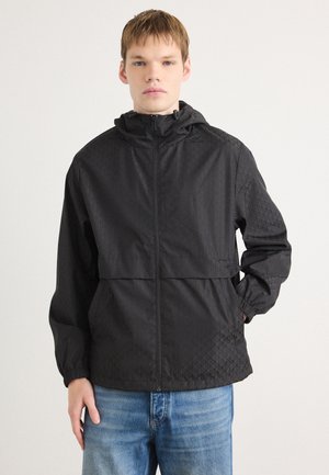 Giacca a vento nera con un motivo geometrico testurizzato; dotata di cappuccio, zip frontale completa e polsi elasticizzati; abbinata a jeans blu.