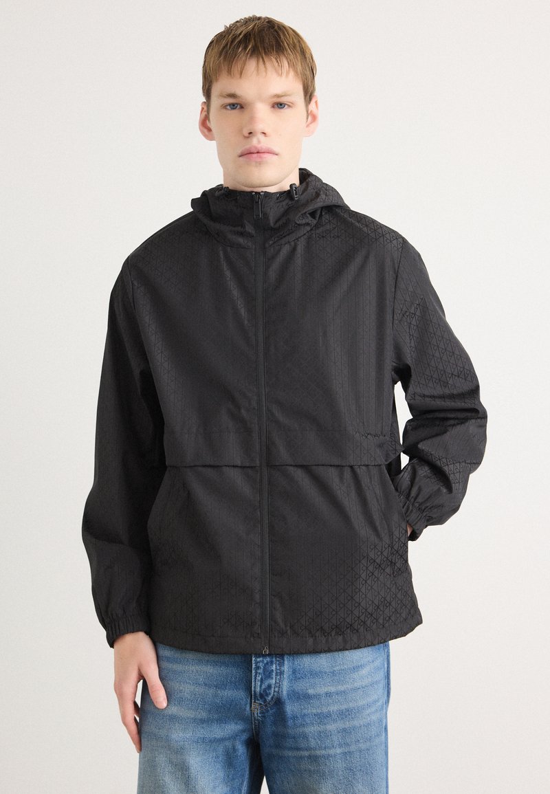 Zwarte windbreaker jas met een gestructureerd, geometrisch patroon; beschikt over een capuchon, volledige ritssluiting aan de voorkant en elastische manchetten; gecombineerd met blauwe jeans.