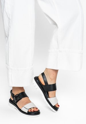 Schwarze und graue Sandalen mit verstellbarer Schnalle, breiten Riemen und weißen Sohlen, kombiniert mit weißen Hosen mit weitem Bein.