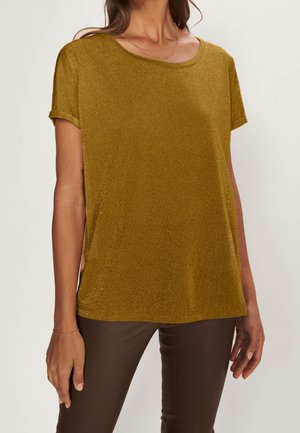 Mujer con una camiseta de manga corta color mostaza con puños remangados y pantalones marrones, mostrada desde el cuello hasta las caderas sobre un fondo liso.