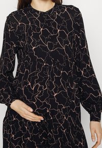 Frau in schwarzem, langärmligem Kleid mit pinkem, abstraktem Linienmuster, eine Hand sanft auf ihrem schwangeren Bauch ruhend.