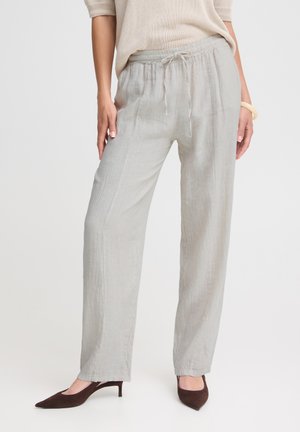 Femme portant un pantalon ample en lin gris clair avec taille à cordon et des chaussures marron à petit talon, debout devant un fond uni.