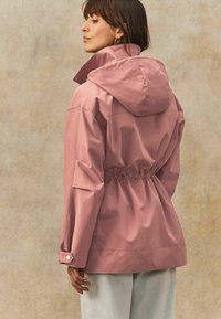Rosa Kapuzenjacke aus glattem, leichtem Material. Mit elastischem Saum und geknöpften Bündchen. Rückansicht, mit lockerer Passform.