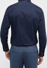 Chemise bleu marine à boutons, manches longues, texture lisse et col classique. Portée avec un pantalon ajusté bleu clair, avec peu de détails.