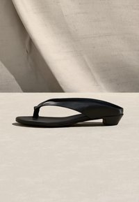 Μαύρο σανδάλι με χαμηλό τακούνι και λεπτά λουράκια, με καμπυλωτή σόλα, τοποθετημένο σε μια ανοιχτή και υφασμάτινη επιφάνεια.