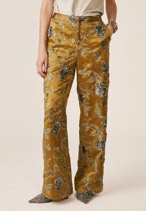 Persona que lleva pantalones de pierna ancha de color mostaza con bordado floral plateado, combinados con zapatos puntiagudos con tachuelas y un top beige.
