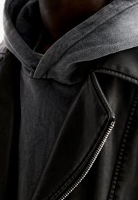 Gros plan sur une personne portant un sweat à capuche gris foncé sous une veste en cuir noir avec une fermeture éclair en argent visible, montrant le cou et le haut de la poitrine.