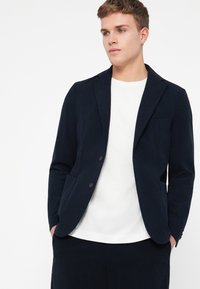 Námornícky modrý blazer vyrobený zo štruktúrovanej tkaniny s dvoma prednými vreckami, zapínaním na jeden gombík a klopami, nosený na bielu košeľu.