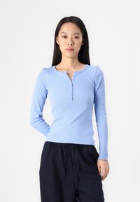 Haut henley à manches longues bleu clair avec un motif en relief texturé, doté d'un col rond et de quatre boutons décoratifs sur le devant.