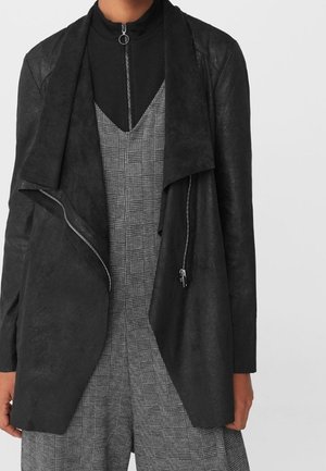 Veste noire en suède ouverte sur une combinaison grise à carreaux avec un top noir à fermeture éclair en dessous, montrée du cou aux hanches.