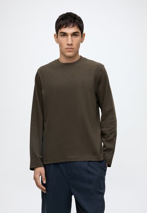Jeune homme aux cheveux courts et foncés portant une chemise à manches longues olive foncé et un pantalon bleu marine, debout devant un fond blanc uni.