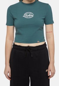 Tyrkysové krátké tričko z měkké bavlny, s bílým logem globu a nápisem "Dickies". Vzhledem ke kombinaci s černými joggery s elastickým pasem.