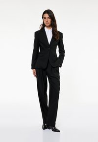 Schwarzer maßgeschneiderter Anzug mit einem Ein-Knopf-Blazer mit Revers, zwei Fronttaschen und hochgeschnittenen, gefältelten Hosen, kombiniert mit einem weißen Hemd.