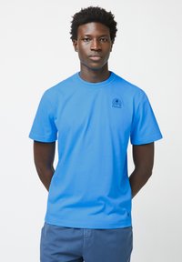 Blaues Baumwoll-T-Shirt mit kurzen Ärmeln und rundem Halsausschnitt. Auf der Vorderseite befindet sich ein kleines dunkelblaues Logo, und der Stoff hat eine glatte, gleichmäßige Textur.