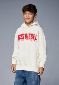 Felpa color crema con un grande logo rosso "DIESEL", tasca anteriore e cappuccio regolabile con cordino. Abbinata a jeans in denim blu.