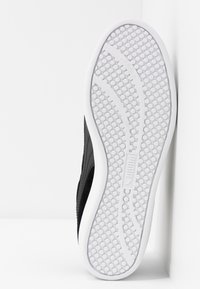 Semelle de chaussure avec motif de grip circulaire et lignes de trace courbées, semelle extérieure blanche avec partie supérieure noire partiellement visible, debout verticalement contre une surface blanche.