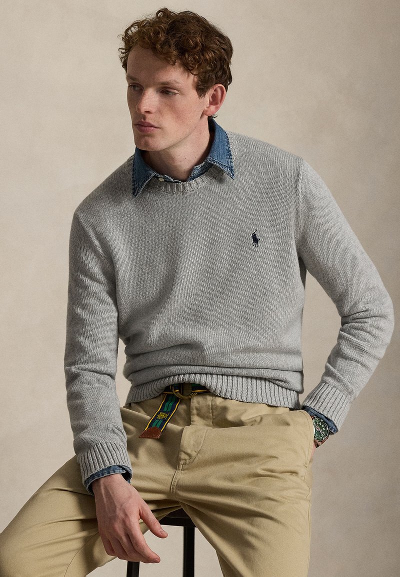 Jonge man met krullend haar, die een grijze trui draagt over een blauwe spijkerbroekoverhemd en beige broek, zittend op een kruk tegen een effen achtergrond.