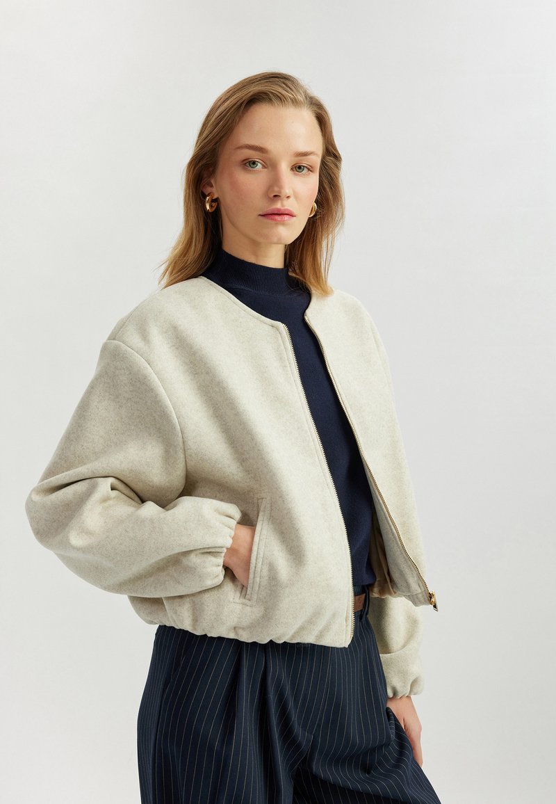 Bomber jacket spesso di colore grigio chiaro, con zip anteriore, polsini elasticizzati e colletto arrotondato. Indossato sopra un dolcevita blu navy, abbinato a pantaloni a righe.