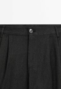 Pantalon gris foncé taille haute avec bouton devant et passants pour ceinture, présentant des plis et un motif de tissu texturé.