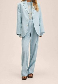 Costume bleu clair sur mesure avec une veste croisée et un pantalon à jambes larges. Tissu lisse, gros boutons et manches évasées créent un look contemporain.