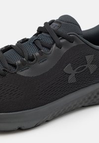 Sort atletisk sko med en tekstureret mesh-overdel, syntetiske detaljer og en gummisål, med sorte snørebånd og et Under Armour-logo.