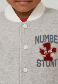 Grijze felt jas met witte ribbelkraag. Bevat lichte knopen en geborduurde details: rode en blauwe tekst die "NUMMER 1 SLVR STUNT" leest.