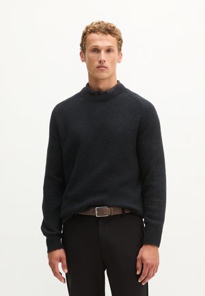 Jonge man met krullend haar, draagt een donker gestructureerde trui over een overhemd met kraag en zwarte broek met een bruine riem, staand voor een effen achtergrond.