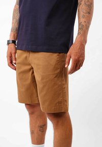 Shorts en coton marron avec une coupe décontractée, dotés de poches latérales et de détails cousus, portés avec un t-shirt bleu foncé et des chaussettes blanches.