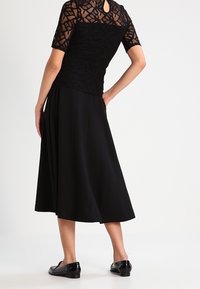 Kvinde iført en kortærmet top i sort blonde og en sort midi-nederdel, med sorte blanke loafers, stående mod en ensfarvet hvid baggrund.