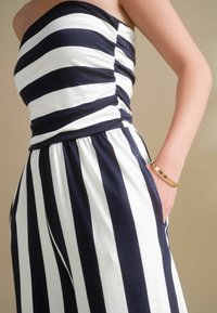Randig jumpsuit i marinblå och vit färg, figursydd bodice med rynkade detaljer, elastisk midja och sidfickor. Guldarmband synligt.
