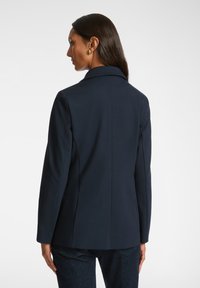 Blazer navy con una vestibilità su misura, rever a punta e maniche lunghe. Tessuto liscio con dettagli minimi, senza motivi o accessori visibili.