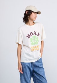 Cream-colored t-shirt with "ROXY" and a green flower graphic. Paired with loose blue denim jeans and a beige cap. Soft fabric, relaxed fit.