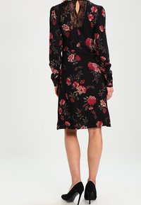 Vestido negro con estampado floral que presenta mangas largas, un detalle de encaje en la parte posterior y un dobladillo a la altura de la rodilla. Acentuado con flores rojas y crema. Tacones altos negros.