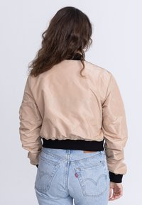 Korte beige bomberjack met een glanzende textuur en zwart geribbeld manchet. Gedragen over lichtblauwe denim jeans met zichtbare achterzakken.