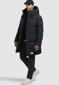 Piumino nero scuro con cappuccio, dotato di chiusura con zip e dettagli in bottone, indossato sopra una maglietta nera e jeans neri, abbinato a sneakers bianche.