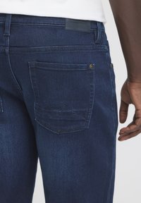 Tumesinised teksapüksid, millel on tagatasku ševroonõmbluse mustriga ja vöökohast ülal nahast silt.