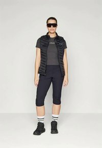 Μαύρο γιλέκο με επένδυση πάνω από γκρι μπλουζάκι, συνδυασμένο με σκούρες κολάν, ριγέ κάλτσες και μαύρες μπότες. Αξεσουάρ περιλαμβάνουν μαύρα γυαλιά ηλίου.