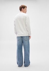 Λευκό φούτερ με μακριά μανίκια και στρογγυλή λαιμόκοψη, συνδυασμένο με ανοιχτό μπλε, φαρδιά τζιν παντελόνια με πίσω τσέπες.