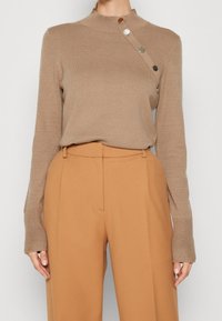 Brauner Strickpullover mit hohem Kragen, der an der Schulter eine diagonale Reihe von metallischen Knöpfen aufweist, kombiniert mit beige hochgeschnittenen Hosen.