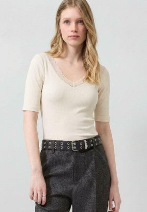 Jonge vrouw met blond haar draagt een crème ribgebreide top met kanten randje en donkere hoge taillebroeken met een zwarte metalen ring riem.