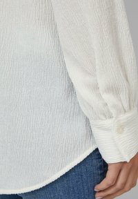 Witte textuur blouse met subtiele verticale strepen, lange mouwen, elastische cuffs en een afgeronde zoom. Gecombineerd met blauwe jeans.