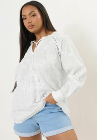Blusa bianca ricamata con scollatura a V e dettaglio con fiocco, abbinata a pantaloni corti in denim azzurro. La camicia presenta intricati motivi floreali.