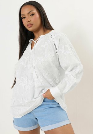 White embroidered blouse with a V-neckline and tie detail, paired with light blue denim shorts. The shirt features intricate floral patterns.