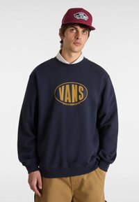 Sudadera azul marino con un gran logo dorado de "VANS" en el centro. La tela parece suave, con un cuello redondo y puños y dobladillo de canalé.