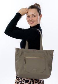 Gray textured tote bag with a front zipper pocket, dual handles, and gold hardware accents, held by a person wearing a black top.
