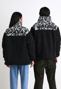 Black fleece jackets with patterned fabric on the shoulders; one has a high collar. Background is plain white. One partner wears light blue jeans, the other wears olive corduroy pants.