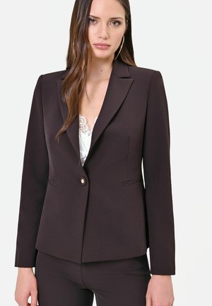 Femme portant un blazer marron foncé ajusté sur un top en dentelle blanche, avec de longs cheveux lisses et de grandes boucles d'oreilles.
