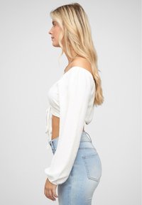 Bílý zkrácený top s rukávy odkrytou ramenou a detailem vázání vpředu, spárovaný s světle modrými vysokými džíny s hladkou texturou.