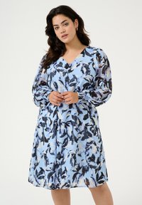 Lyseblå kjole med et blomsterprint i sort og hvid, V-hals, lange gennemsigtige ærmer og en indsnævret talje.